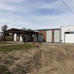 Nowoczesny budynek z elementami drewnianymi, plac zabaw z huśtawką i karuzelą, widoczne panele słoneczne na dachu, teren wokół w trakcie zagospodarowania.