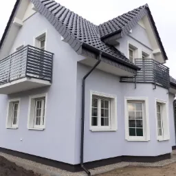 Narożne ujęcie świeżo wykończonego domu w odcieniach szarości z czarną dachówką i balkonami z metalowymi barierkami, widoczne fragmenty ogrodu w stanie surowym.