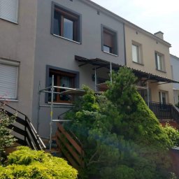 BOA Rafał Duś - Szara elewacja domu jednorodzinnego w trakcie prac, widoczne rusztowania i stolarka okienna z brązowymi ramami, bujna zieleń w ogrodzie.