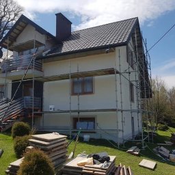 BOA Rafał Duś - Dom jednorodzinny w trakcie prac elewacyjnych, z widocznymi rusztowaniami, częściowo ocieploną ścianą, paletami z materiałami budowlanymi na trawniku i błękitnym niebem w tle.