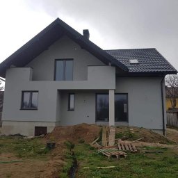 BOA Rafał Duś - Nowoczesny dom jednorodzinny z szarą elewacją, czarną dachówką i stolarką okienną, widoczny w pochmurny dzień, z przygotowanym terenem wokół budynku.