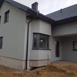 BOA Rafał Duś - Dom jednorodzinny po tynkowaniu elewacji na szaro, z widocznymi czarnymi oknami i dachem z dachówki, teren wokół domu częściowo uporządkowany.