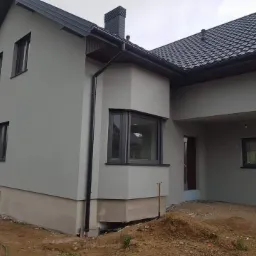 Dom jednorodzinny po tynkowaniu elewacji na szaro, z widocznymi czarnymi oknami i dachem z dachówki, teren wokół domu częściowo uporządkowany.