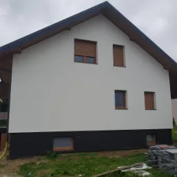 Dwukolorowa elewacja domu jednorodzinnego z brązowymi roletami okiennymi, widok z boku na tle pochmurnego nieba, z ułożonymi płytkami chodnikowymi na pierwszym planie.