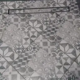 Ułożone pod kątem płytki podłogowe w łazience z wzorem patchworkowym w odcieniach szarości, z liniowym odpływem w rogu. Widoczne ciemnoszare płytki ścienne.