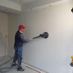 BOA Rafał Duś - Mężczyzna w czerwonej czapce szlifuje ścianę wewnątrz budynku za pomocą szlifierki z odkurzaczem; widoczny również statyw z oświetleniem.