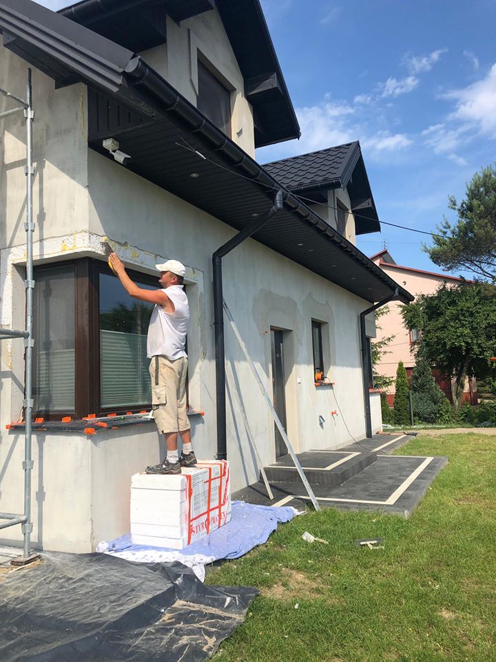Pracownik w białej koszulce i kapeluszu, stojąc na styropianie, wygładza zaprawę wokół okna na elewacji domu jednorodzinnego przed ociepleniem.