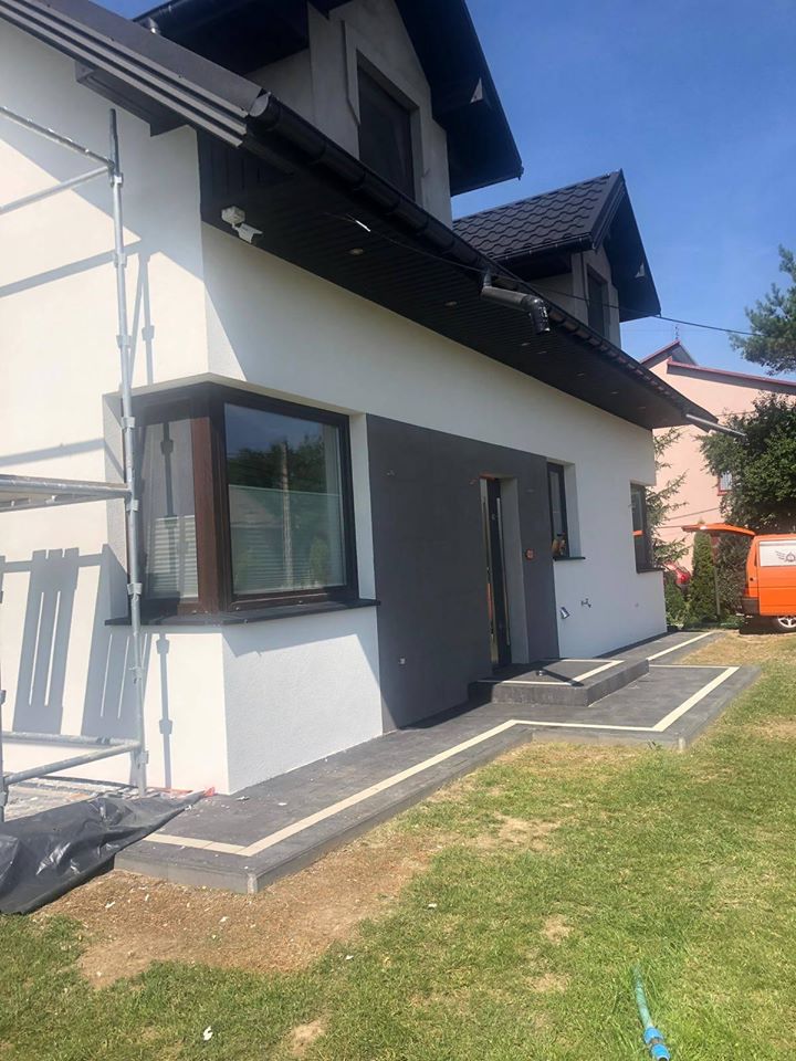 Dwukondygnacyjny dom z elewacją w odcieniach bieli i szarości, z czarną stolarką okienną i dachem; widoczne rusztowania i nowa ścieżka z kostki brukowej.