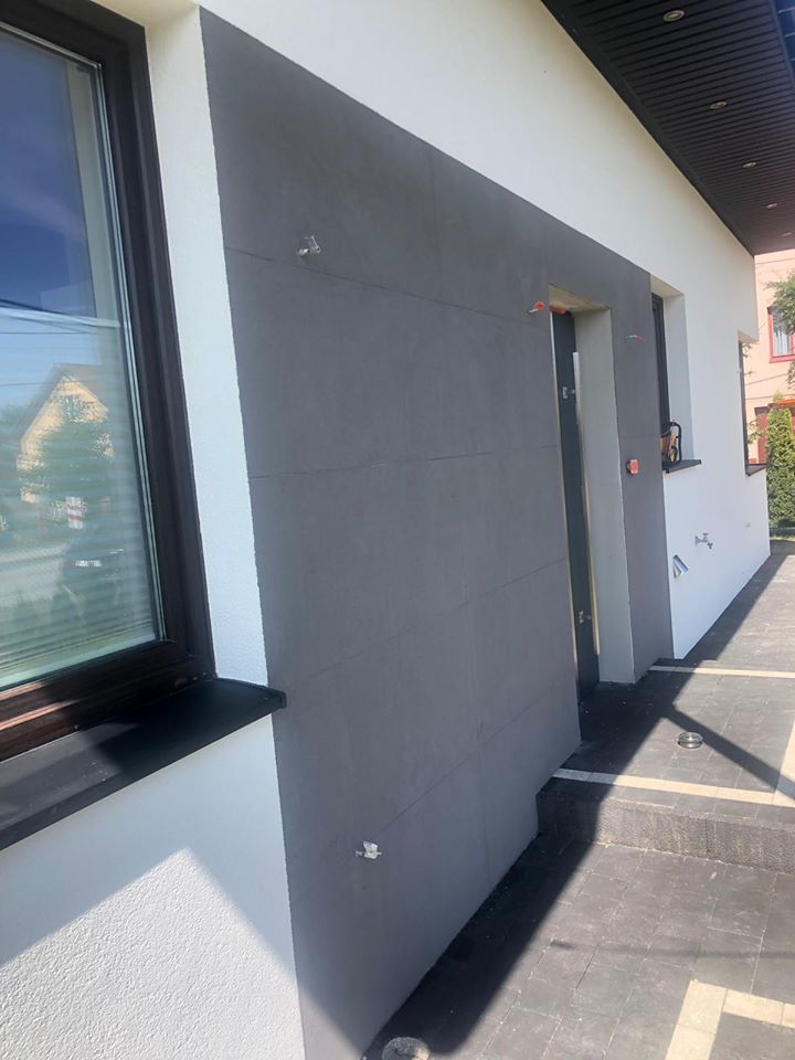 Szara elewacja budynku w trakcie prac, widoczne mocowania izolacji, okna z ciemnymi ramami i fragment chodnika z kostki brukowej.