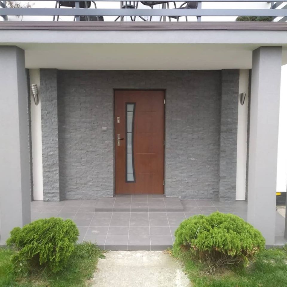 Wejście do domu z brązowymi drzwiami i szarą elewacją z kamienia dekoracyjnego, oświetlone kinkietami, z szarą podłogą wyłożoną płytkami i ozdobnymi krzewami po bokach.