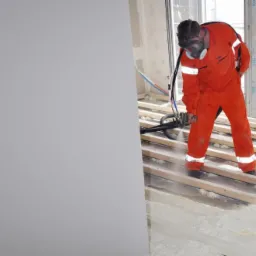 Pracownik w kombinezonie i masce natryskuje pianę izolacyjną na drewnianą podłogę w jasnym wnętrzu. Widoczne okno i fragment ściany.