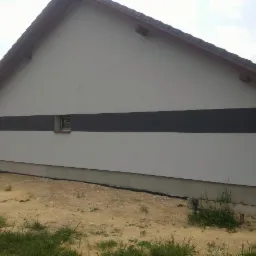 Wykończona elewacja domu z szarą, poziomą linią. Widoczne okna i fragment dachu. Dom stoi na działce z trawą i piaskiem.