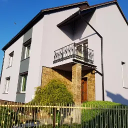 Dwukondygnacyjny dom z jasnoszarą elewacją, kamiennym wykończeniem kolumn balkonu i fragmentem szarej ściany bocznej, otoczony metalowym ogrodzeniem.