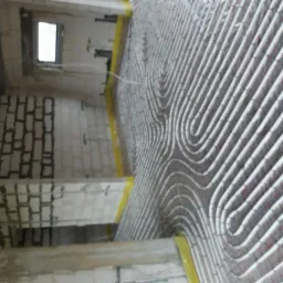 Układanie rur ogrzewania podłogowego w nowym budynku z surowymi ścianami z betonu komórkowego i oknem. Rury ułożone spiralnie, zabezpieczone żółtą taśmą na obrzeżach.