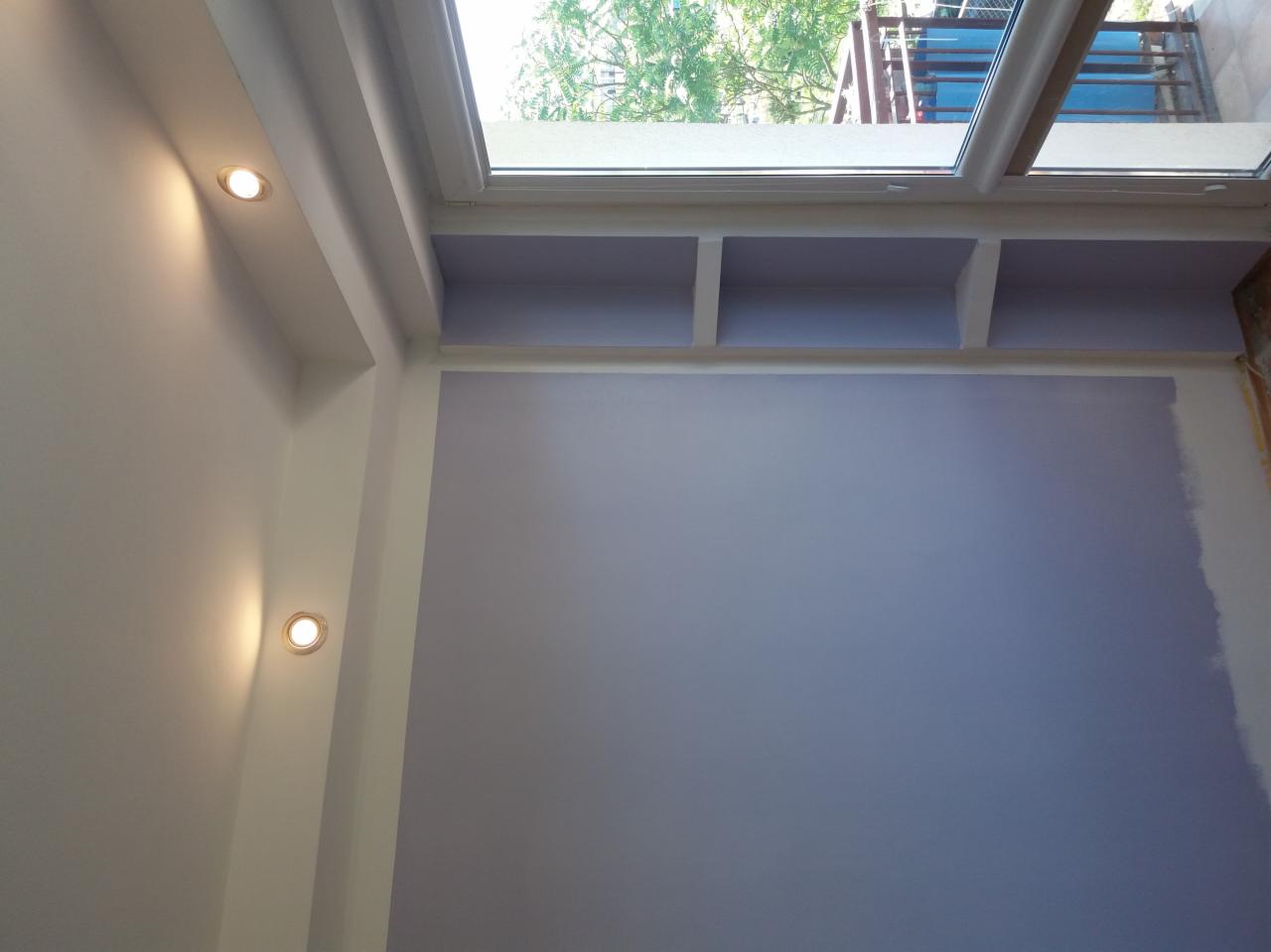 Pomieszczenie w trakcie malowania na fioletowo, widoczne wnęki pod sufitem i okno dachowe z widokiem na drzewa, dwie lampy punktowe na ścianie.