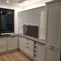 Jasna kuchnia z drewnianą podłogą i oknem. Szafki górne z przeszklonymi frontami. Dolne szafki z szufladami i miejscem na wino. Blat w kolorze szarości.