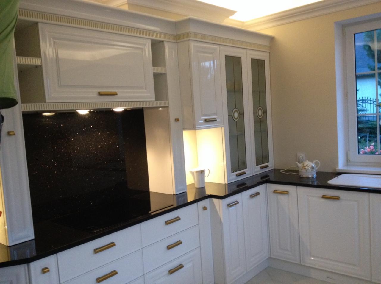 Biała kuchnia z czarnym blatem i złotymi uchwytami, oświetlona, z widocznym oknem i czajnikiem. Klasyczny styl z dekoracyjnymi elementami.