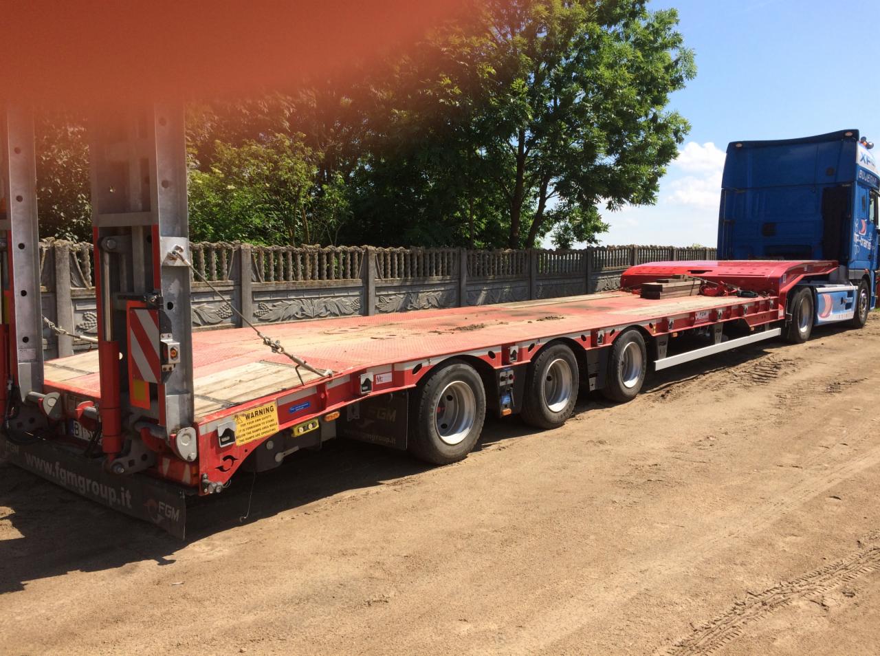 Czerwona naczepa niskopodwoziowa z niebieskim ciągnikiem siodłowym na piaszczystym placu, widoczny fragment betonowego ogrodzenia w tle.