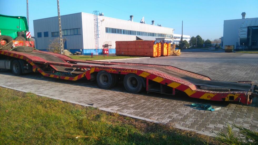 Czerwono-żółta naczepa niskopodwoziowa na placu przemysłowym z budynkami w tle, widoczne opony na kabinie ciągnika.