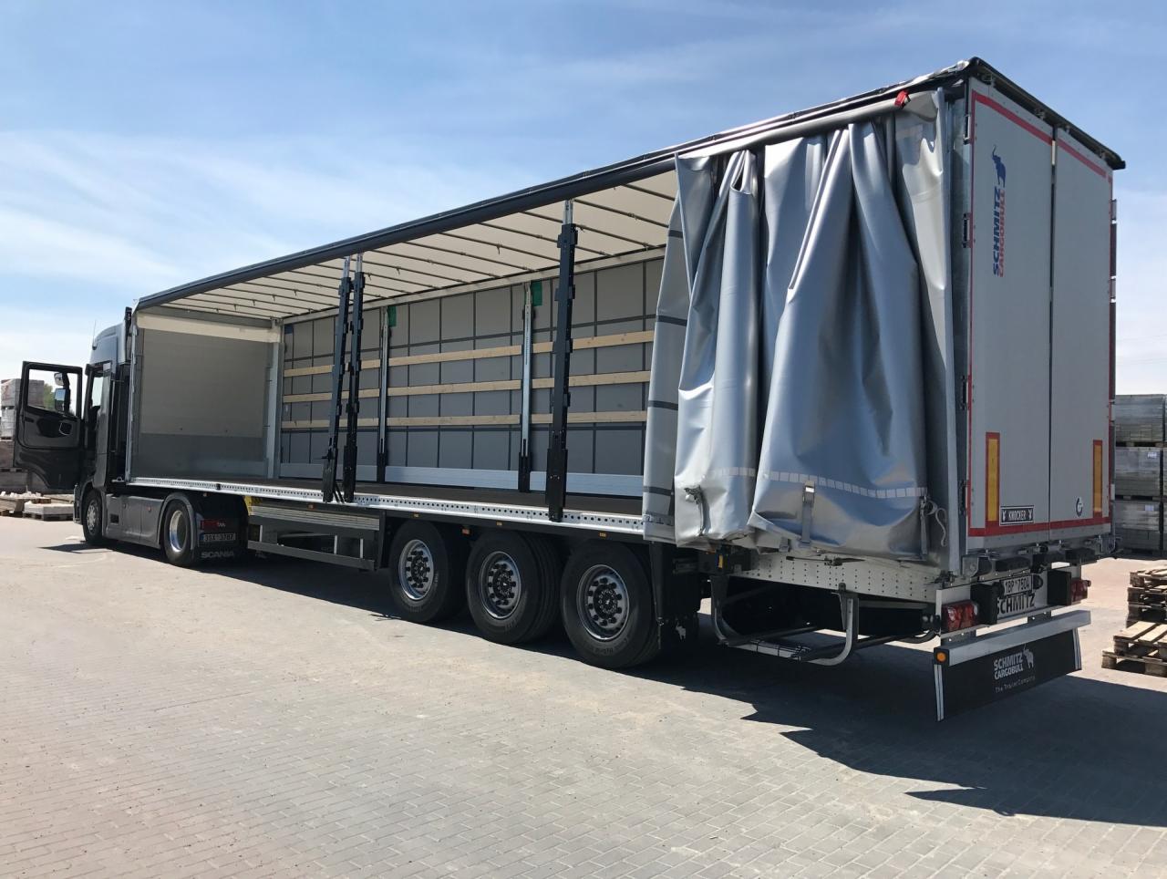 Ciężarówka marki Scania z otwartą naczepą Schmitz Cargobull, z widocznymi zabezpieczeniami ładunku, stojąca na betonowym placu w słoneczny dzień.