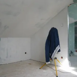 Pokój na poddaszu w trakcie prac wykończeniowych; ściany i sufit częściowo pokryte gładzią, lampa robocza oświetla wnętrze. Widoczne profile.