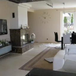 Jasne wnętrze salonu z marmurowym kominkiem, dużym zegarem ściennym i wyjściem na ogród przez przeszklone drzwi. Widoczna część białej sofy.