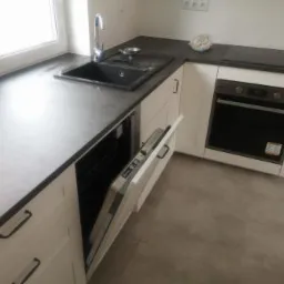 Fragment nowoczesnej kuchni z białymi szafkami z frezowanymi frontami, ciemnym blatem, czarnym zlewozmywakiem i otwartą zmywarką, widoczne okno i płyta grzewcza.