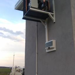 Naprawa Sprzętu AGD i Urządzeń Chłodniczych Karolkowski Leszek - Zewnętrzna jednostka chłodnicza zamontowana na ścianie budynku, widoczne miedziane rurki, biała obudowa i wsporniki mocujące. W tle częściowo widoczny samochód dostawczy.