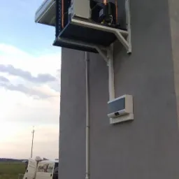 Zewnętrzna jednostka chłodnicza zamontowana na ścianie budynku, widoczne miedziane rurki, biała obudowa i wsporniki mocujące. W tle częściowo widoczny samochód dostawczy.