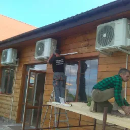 Montaż kilku klimatyzator&oacute;w na drewnianej elewacji budynku, dwaj monterzy na rusztowaniu, jeden w koszulce z napisem branżowym, pudełka klimatyzator&oacute;w Inventor na ziemi.