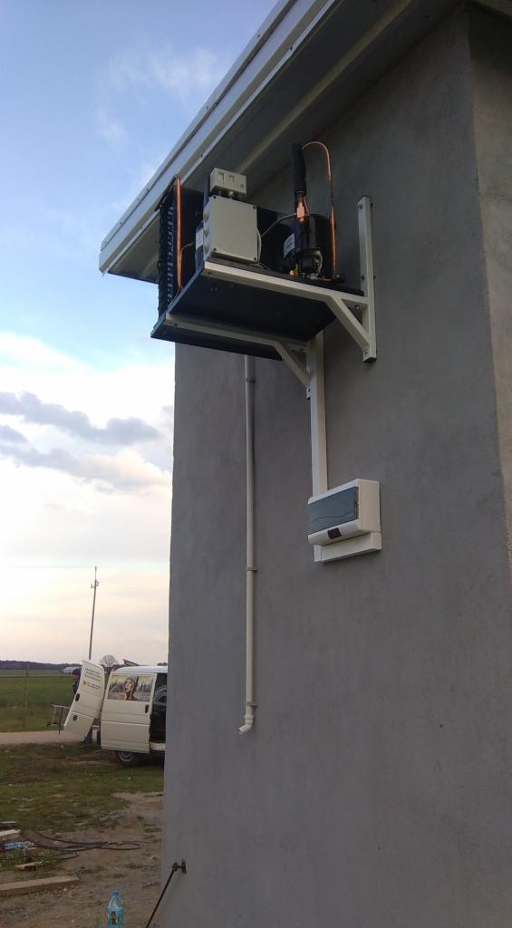 Zewnętrzna jednostka chłodnicza zamontowana na ścianie budynku, widoczne miedziane rurki, biała obudowa i wsporniki mocujące. W tle częściowo widoczny samochód dostawczy.