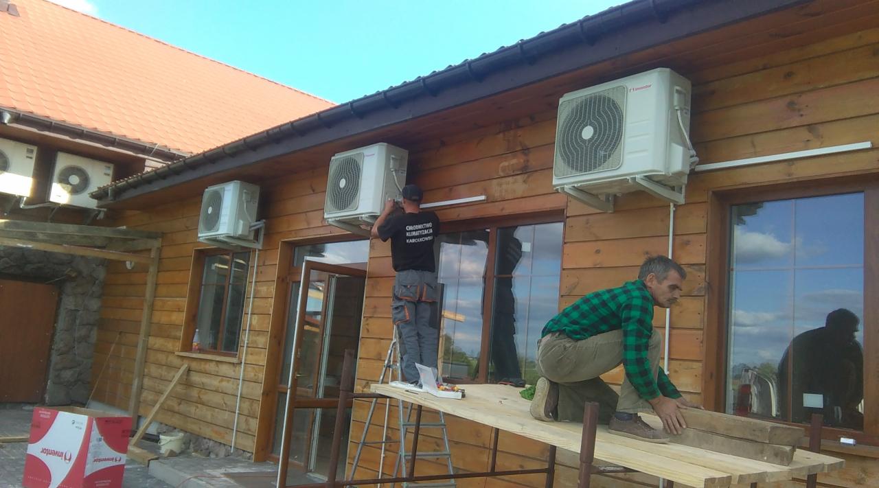 Montaż kilku klimatyzatorów na drewnianej elewacji budynku, dwaj monterzy na rusztowaniu, jeden w koszulce z napisem branżowym, pudełka klimatyzatorów Inventor na ziemi.
