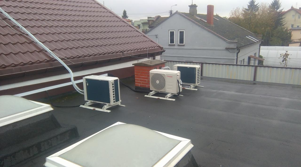 Trzy jednostki zewnętrzne klimatyzacji zamontowane na płaskim dachu budynku, widoczne przewody, w tle budynki mieszkalne i pochmurne niebo.
