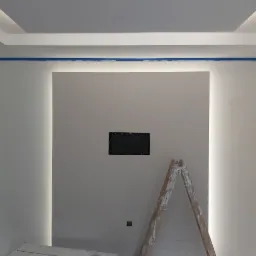 Ściana w trakcie przygotowania do malowania z widoczną niebieską taśmą malarską, gładzią i oświetleniem LED w suficie podwieszanym oraz drabina malarska przy ścianie z otworem na gniazdko.