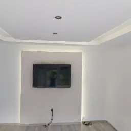 Nowoczesne wnętrze salonu z podwieszanym sufitem z oświetleniem LED, minimalistyczna ściana z telewizorem, jasne ściany i podłoga imitująca drewno.