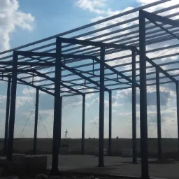 Szkielet konstrukcji z ciemnej stali pod budowę hali na tle błękitnego nieba z chmurami i odległego krajobrazu polnego z widocznymi słupami wysokiego napięcia.