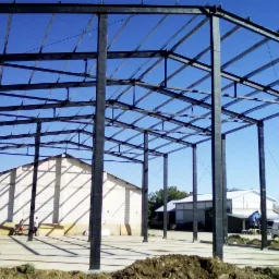 Czarna, stalowa konstrukcja szkieletowa hali przemysłowej w trakcie budowy, widoczne fundamenty, dwóch pracowników w oddali i budynek w tle pod błękitnym niebem.