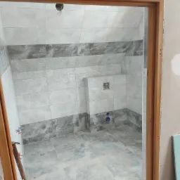 Wnętrze łazienki w trakcie remontu z częściowo ułożonymi szaro-białymi płytkami na ścianach i podłodze, widoczne wystające instalacje wodne, surowa ściana z płytą karton gips oraz drewniana...