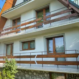 Dwupiętrowy dom z jasną elewacją, drewnianymi oknami i balkonami z drewnianą balustradą o charakterystycznym wzorze, otoczony zielenią.