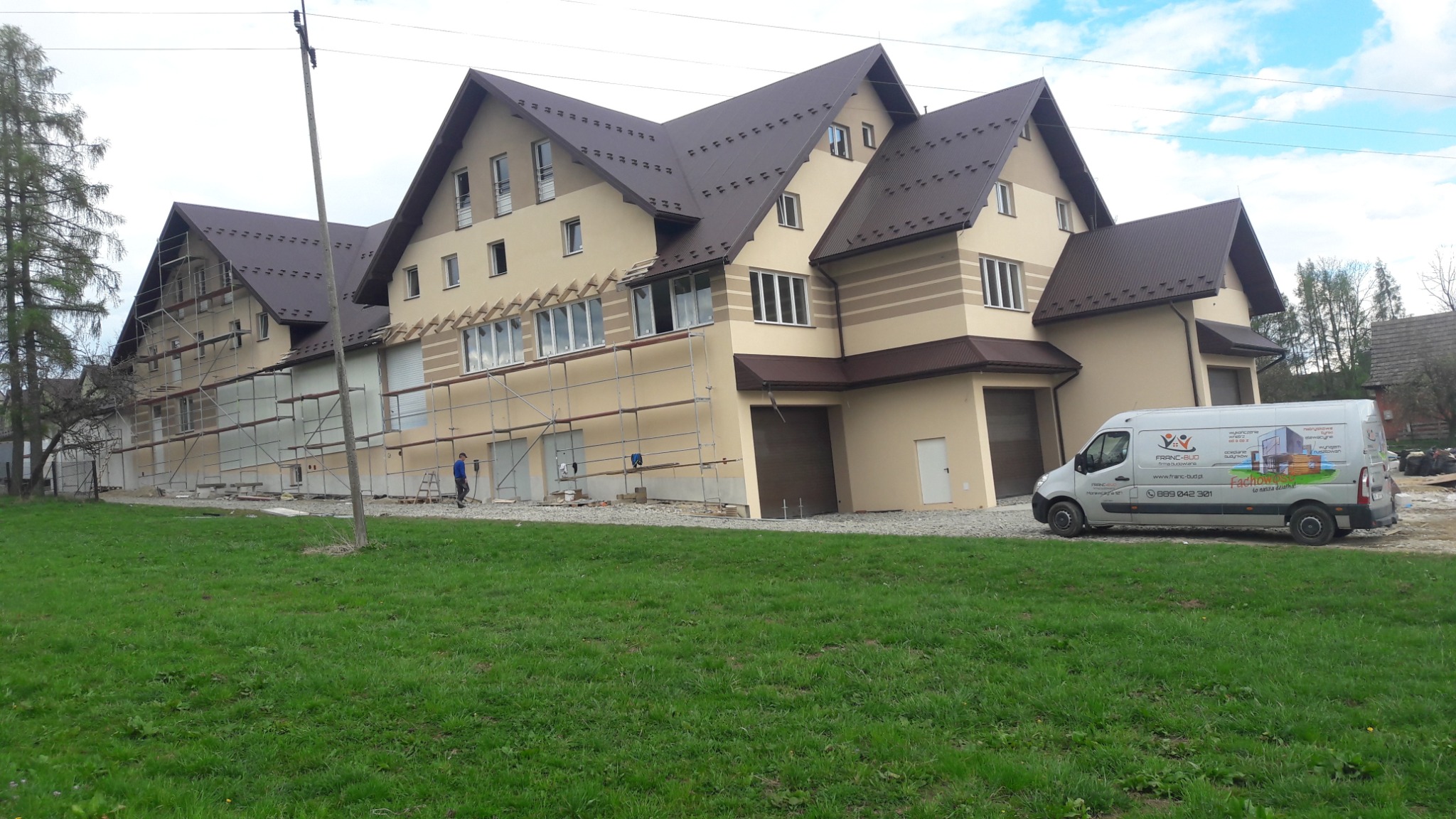 Budynek w Lasku z częściowo wykonaną elewacją, widoczne rusztowania i pracownik w oddali, na pierwszym planie trawnik i firmowy bus z reklamą.