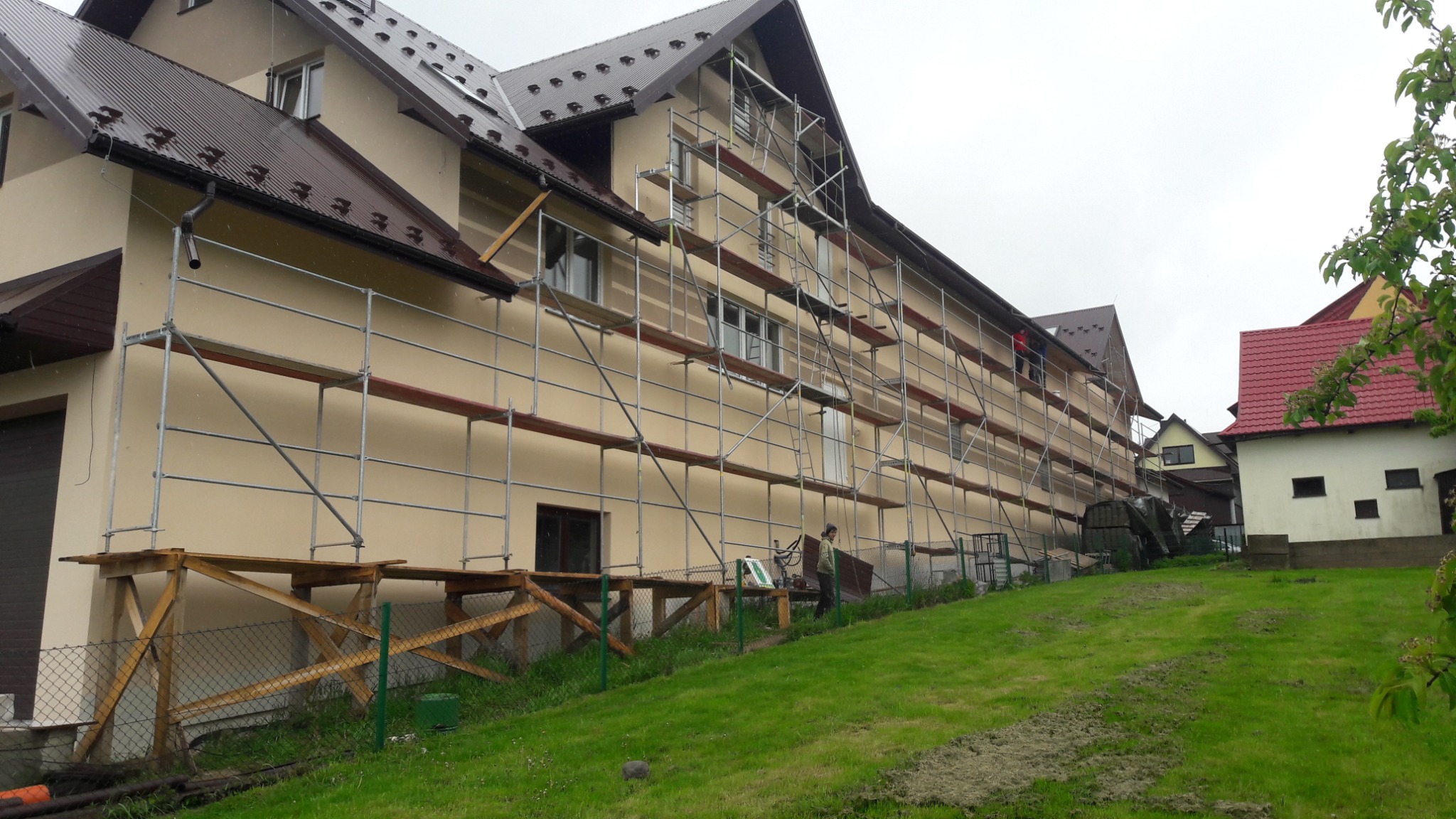 Długi budynek w trakcie prac elewacyjnych, z rozstawionymi rusztowaniami i pracownikiem na górze, widok z zielonego trawnika.