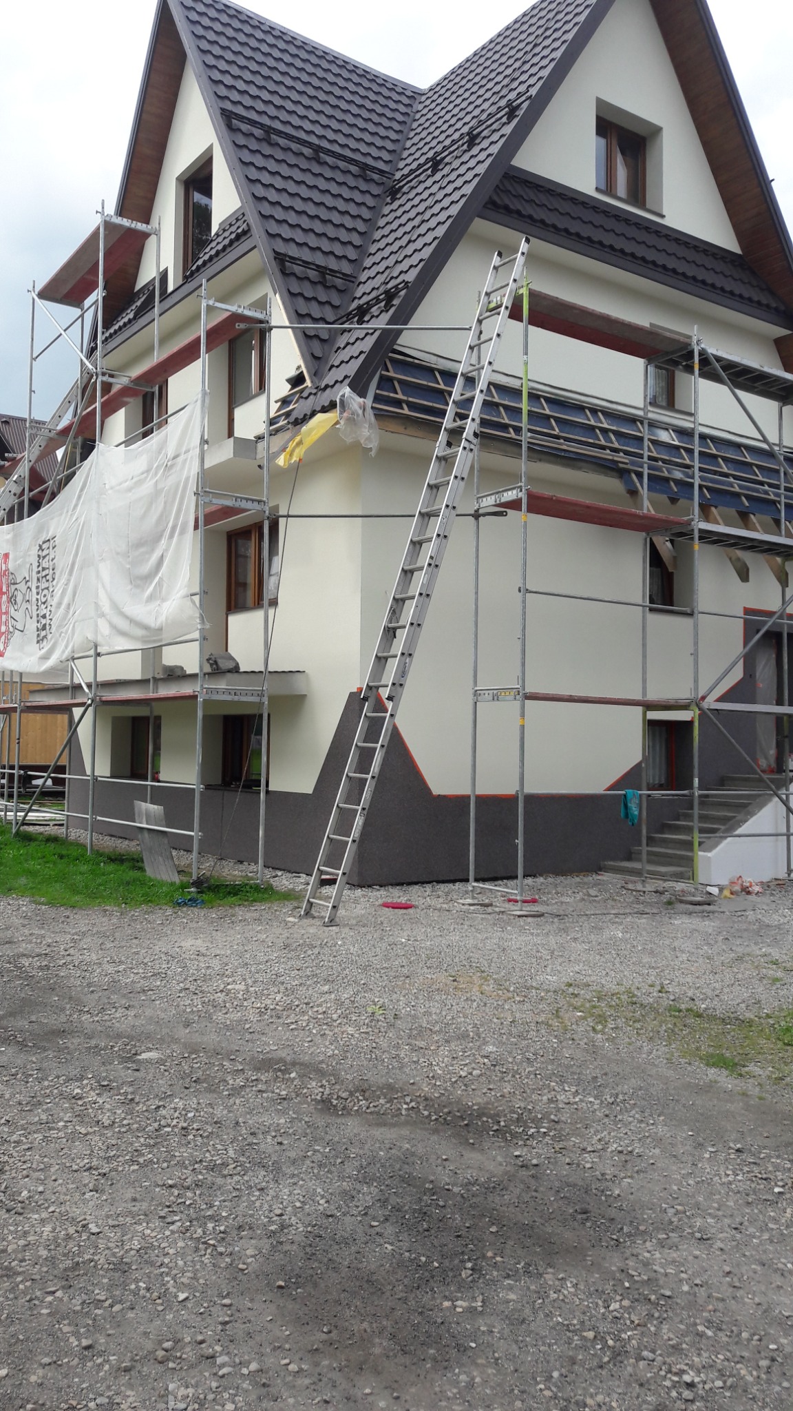 Dom jednorodzinny w trakcie prac elewacyjnych, widoczne rusztowania, drabina oparta o ścianę, częściowo otynkowana elewacja i ocieplony cokół budynku.