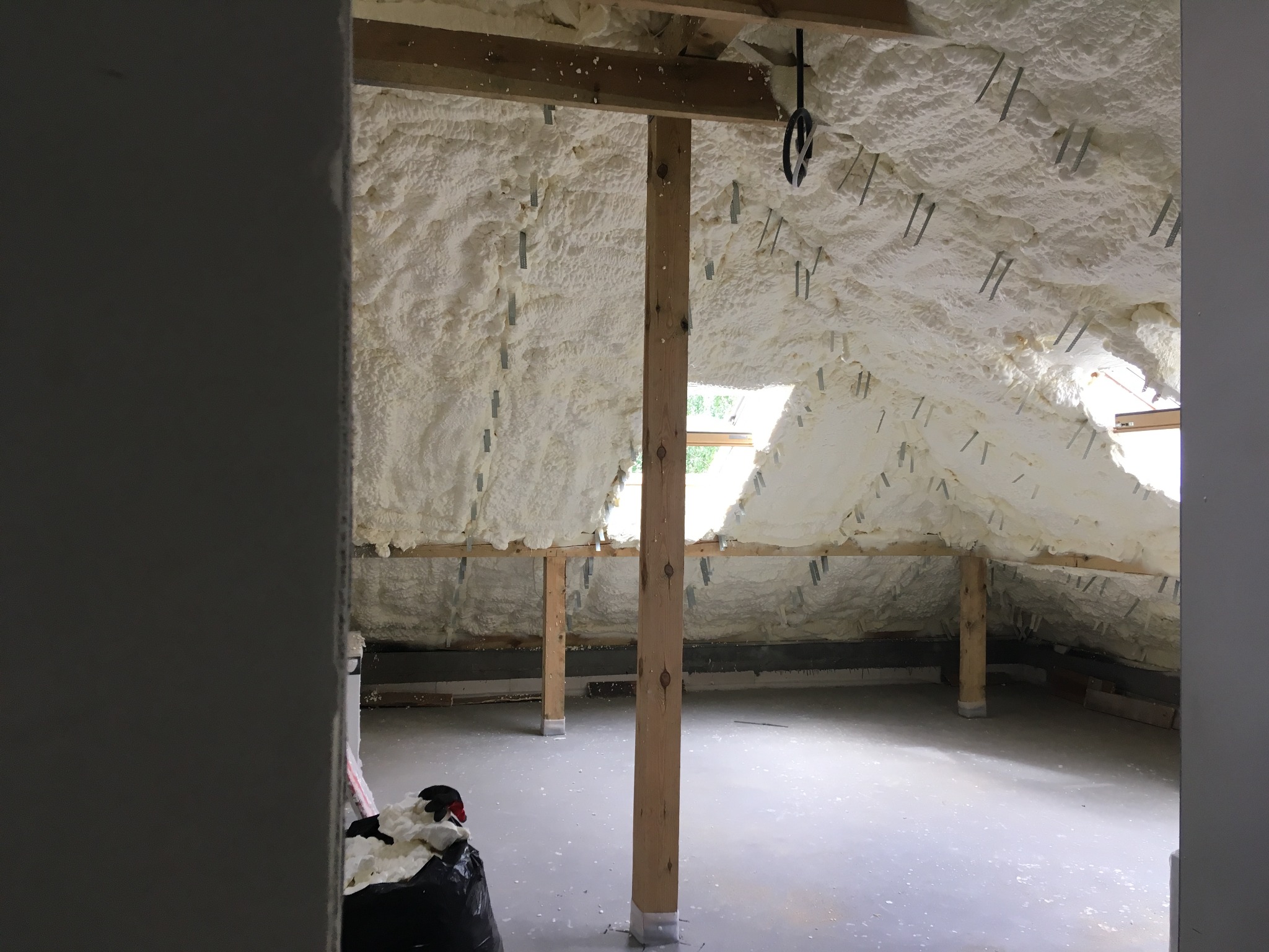 Poddasze w trakcie ocieplania pianą poliuretanową, widoczne drewniane elementy konstrukcyjne i metalowe mocowania, przez okna widać zieleń drzew.