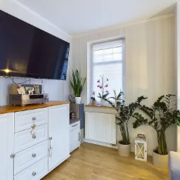 Jasne wnętrze salonu z dużym telewizorem, białą komodą z drewnianym blatem i roślinami doniczkowymi przy oknie. Na komodzie sprzęt audio i dekoracje.