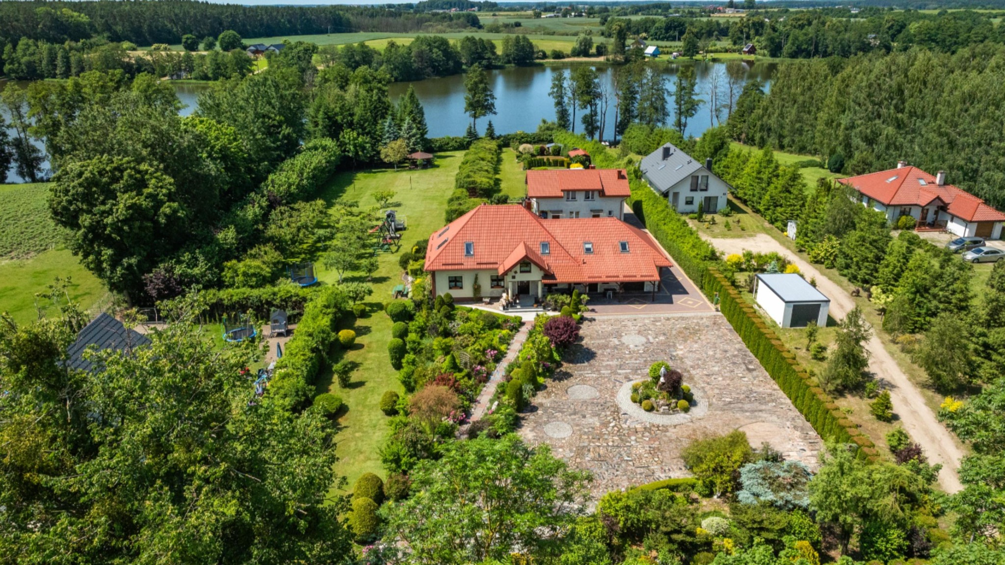 Panoramiczny widok z lotu ptaka na rozległą posiadłość z domem z czerwoną dachówką, otoczoną zielenią, z jeziorem w tle, w słoneczny dzień.