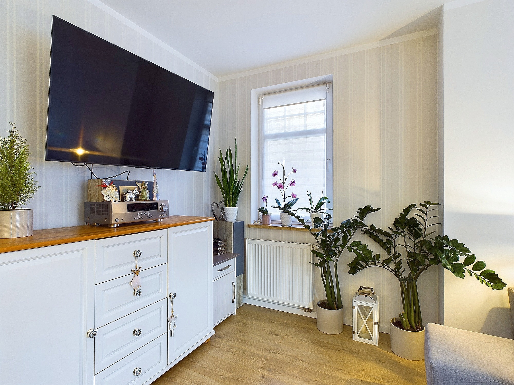 Jasne wnętrze salonu z dużym telewizorem, białą komodą z drewnianym blatem i roślinami doniczkowymi przy oknie. Na komodzie sprzęt audio i dekoracje.