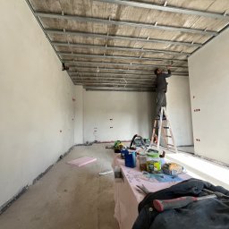 TM Milena Tomczak - Montaż metalowego stelażu pod konstrukcję sufitu w surowym wnętrzu; pracownik na drabinie mocuje elementy, widoczne narzędzia i materiały na stole roboczym, surowe ściany i podłoga w trakcie prac...