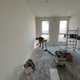 TM Milena Tomczak - Pracownik szlifuje ścianę w nowym mieszkaniu, używając szlifierki z odkurzaczem, podłoga zabezpieczona folią malarską, widoczne narzędzia i drabina.
