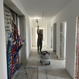 TM Milena Tomczak - Pracownik szlifuje sufit w długim korytarzu podczas prac wykończeniowych, widoczne instalacje hydrauliczne i wentylacyjne, szlifierka na wysięgniku i odkurzacz przemysłowy.