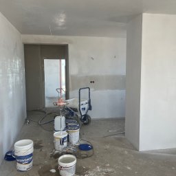 TM Milena Tomczak - Wnętrze w trakcie prac wykończeniowych: świeżo pomalowane ściany na biało, widoczne ślady po wałku i mokra powierzchnia, na podłodze puste i częściowo puste wiadra po farbie, agregat malarski...